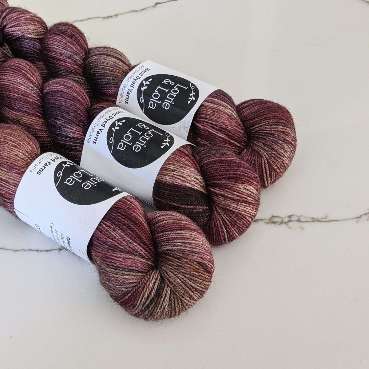 Multiple skeins of purple and burgundy hand-dyed yarn labeled "Louie & Lola" on white marble surface.