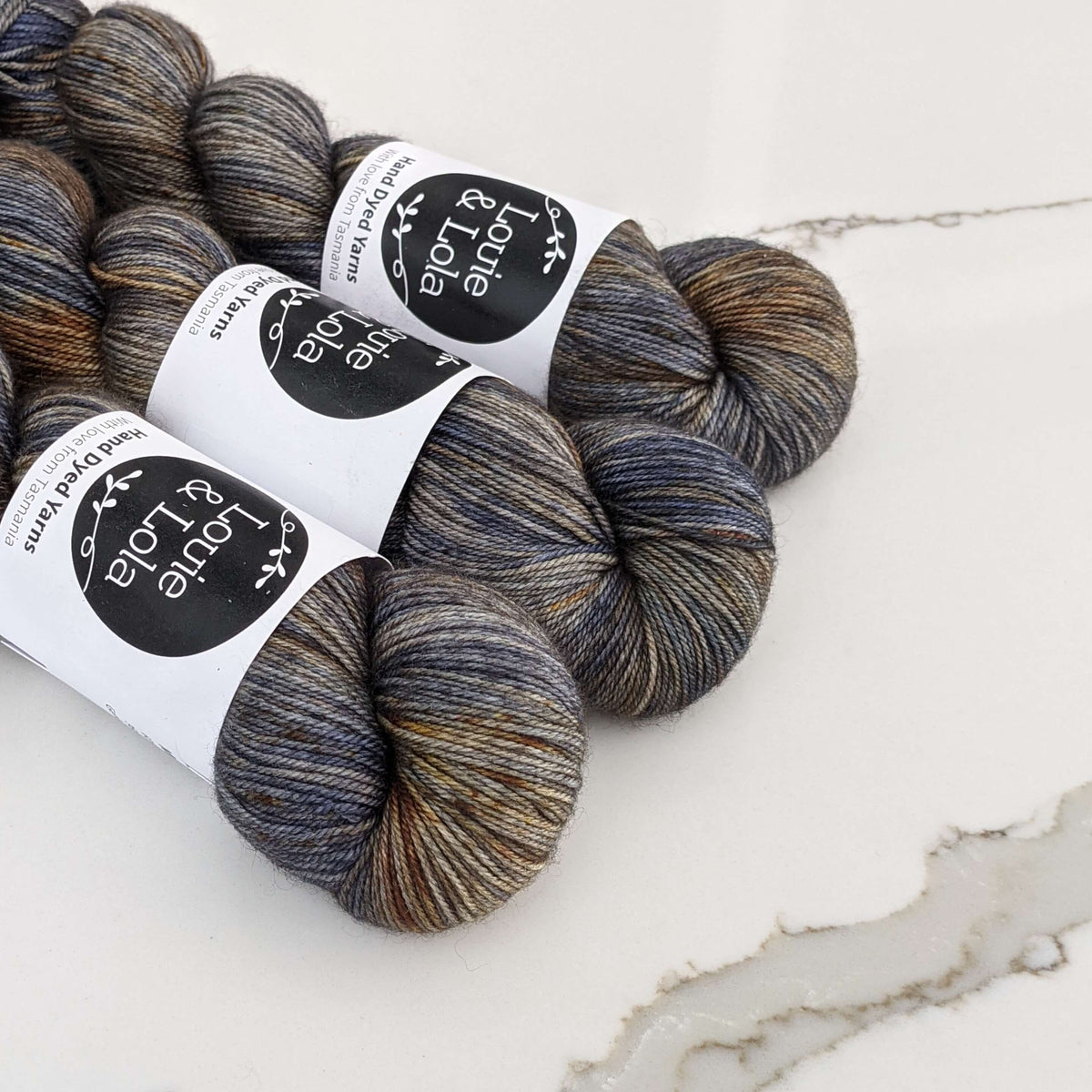 Three skeins of dark grey and brown marbled yarn with labels on a white marble surface.