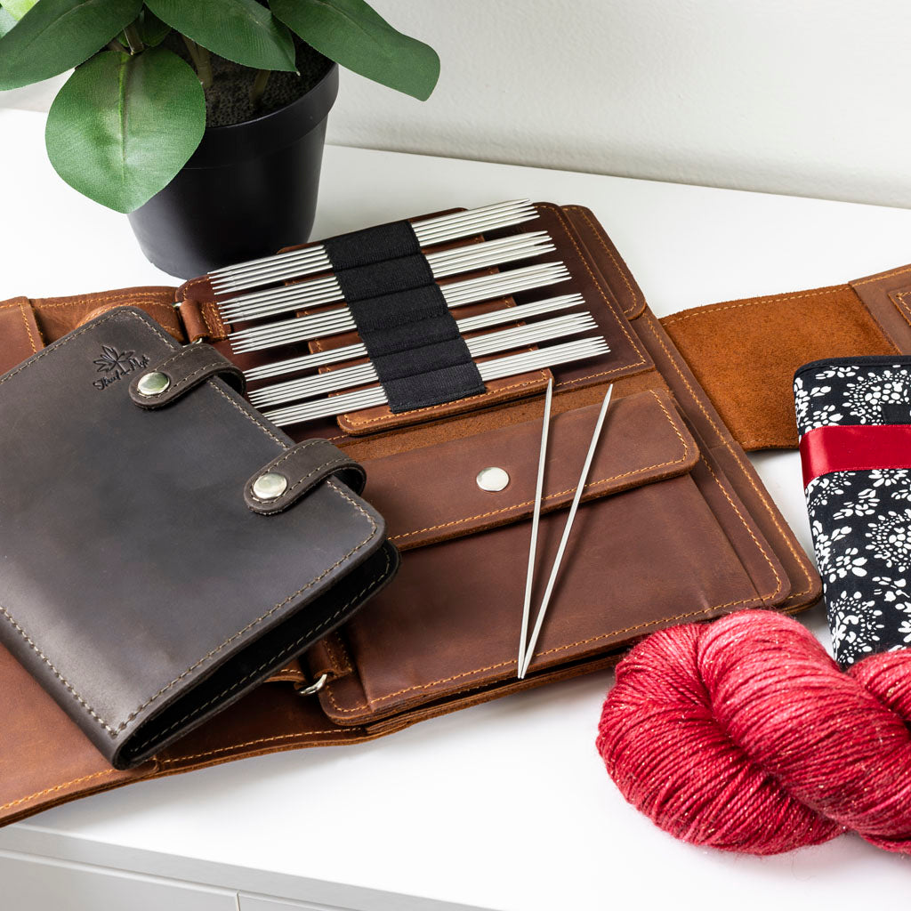 Leather knitting needle case with steel double-pointed needles, red yarn, and a decorative fabric pouch on a white table beside a potted plant.