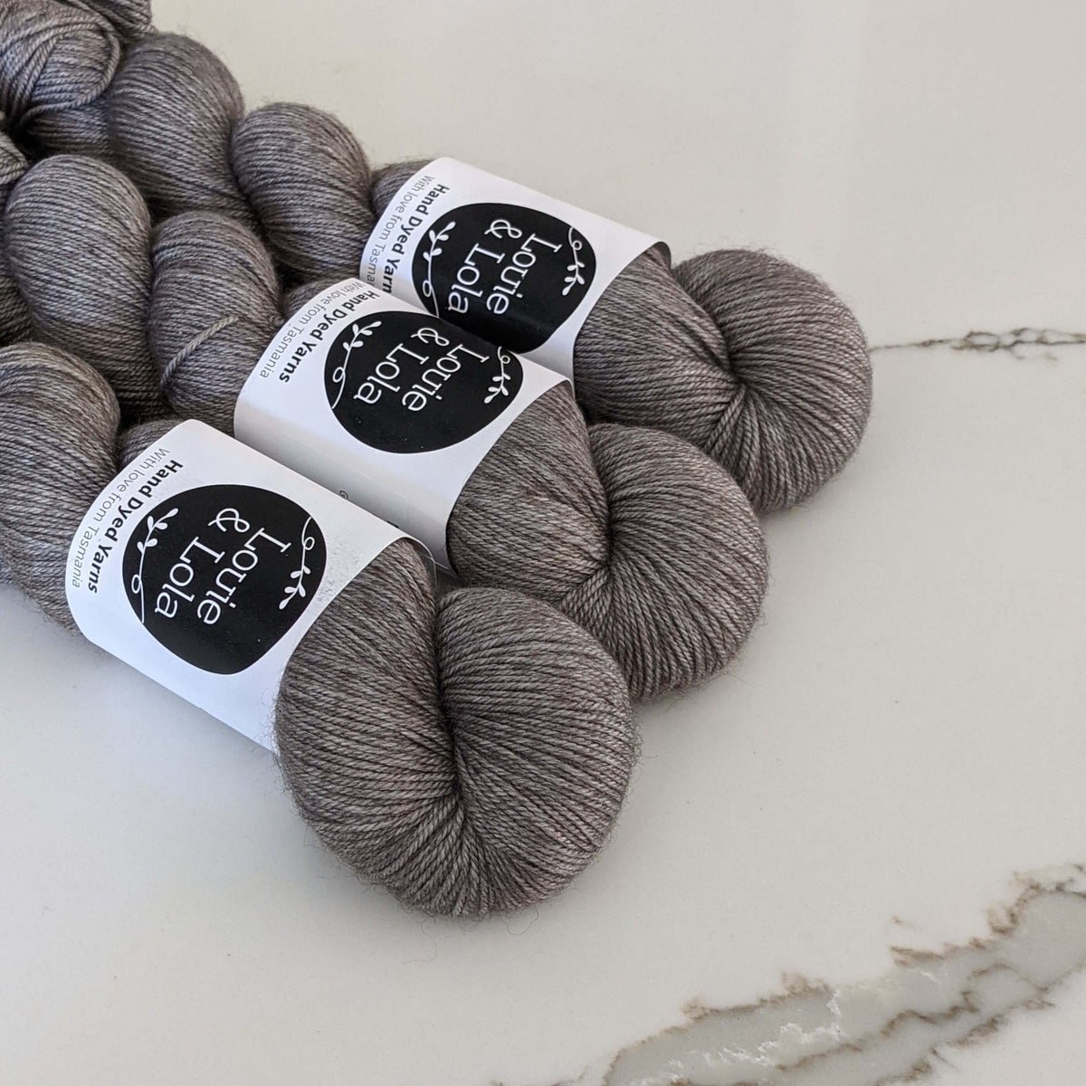 Three skeins of hand-dyed Louie & Lola grey yarn on a marble surface.