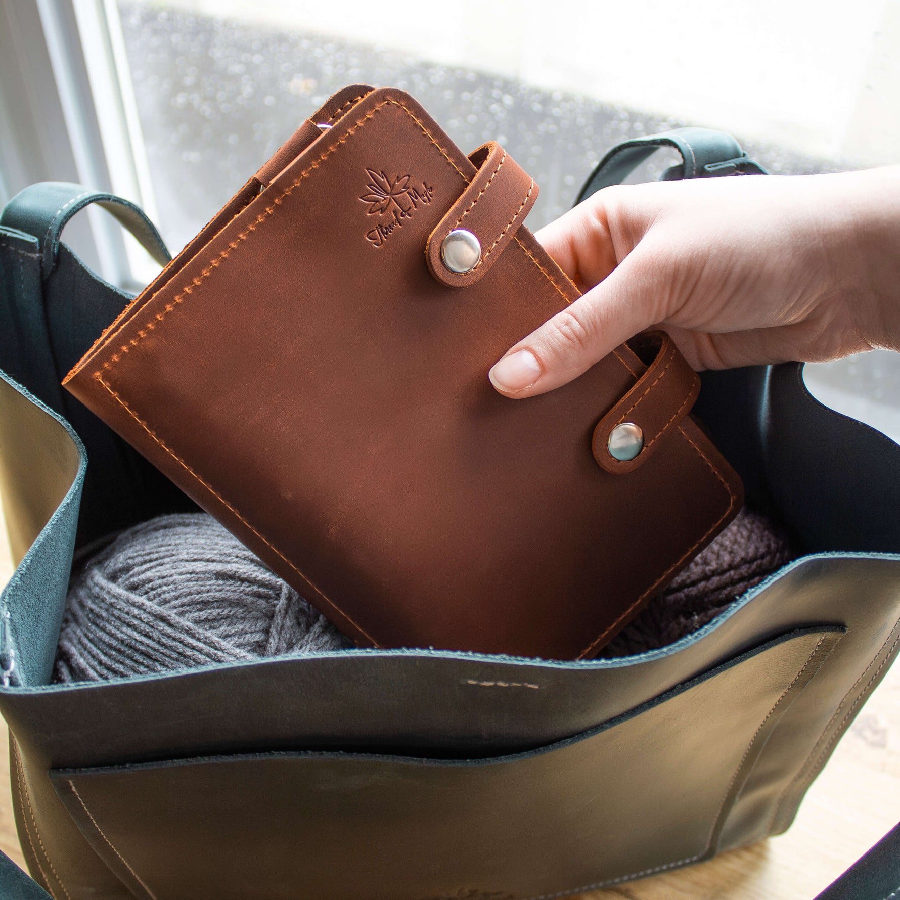 Hand placing a brown leather notebook into a tote bag containing gray yarn near a window.