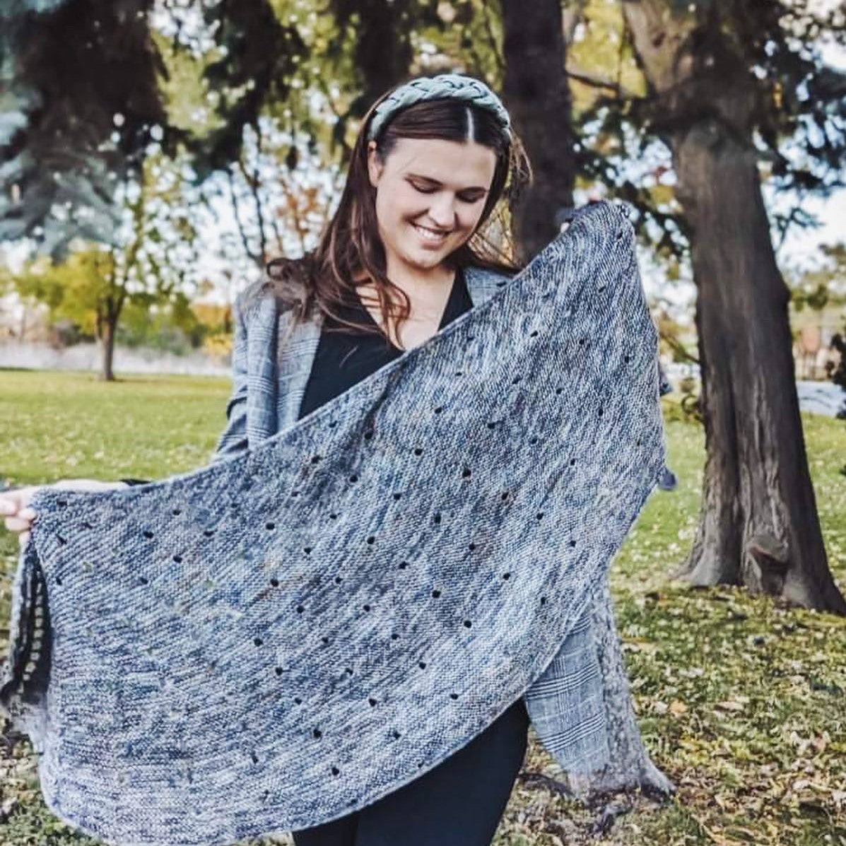 A woman smiling while holding a knitted gray shawl with a lacy pattern in a park setting with trees and grass.