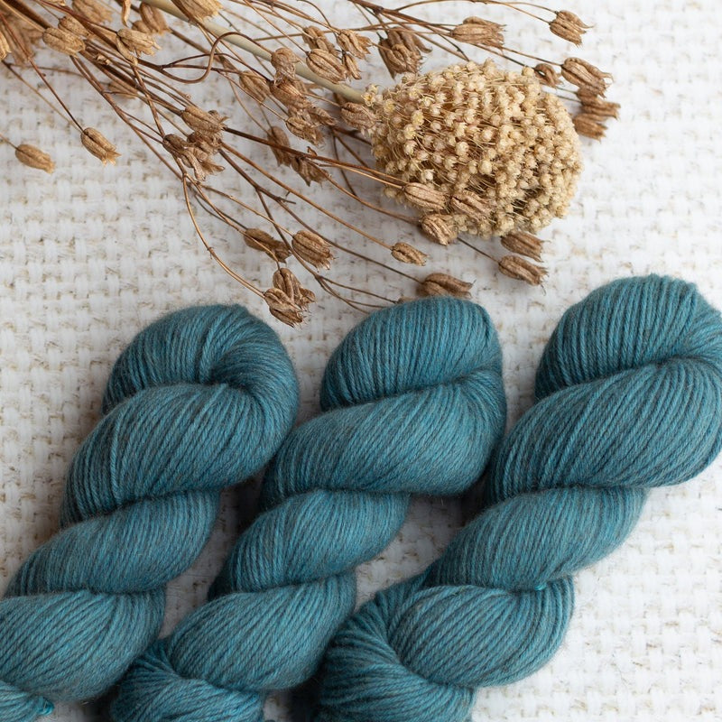 Three skeins of blue yarn placed on a textured white background with dried floral arrangement.