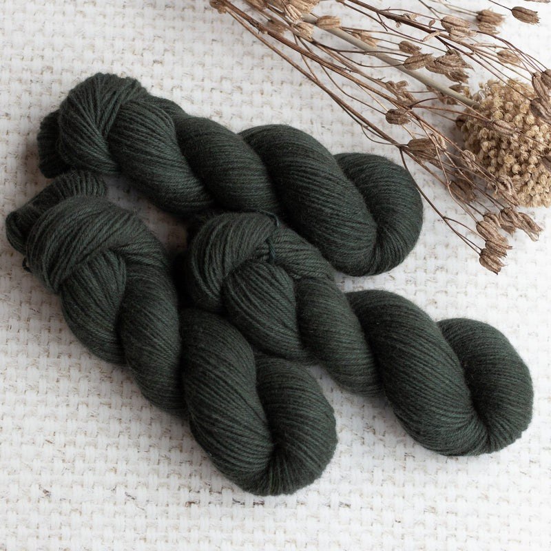 Dark green yarn skeins arranged on a woven fabric surface with dried plant stems in the background.