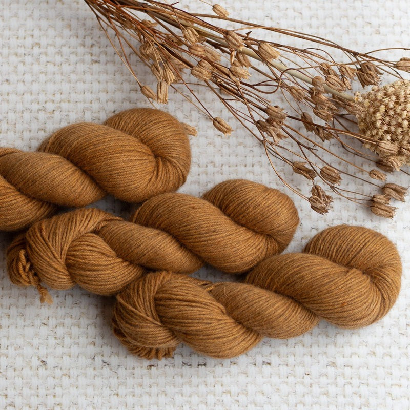 Brown wool yarn skeins on textured white fabric with dried floral sprigs.