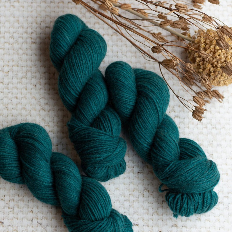 Three skeins of dark teal yarn on a textured surface with dried flowers nearby.