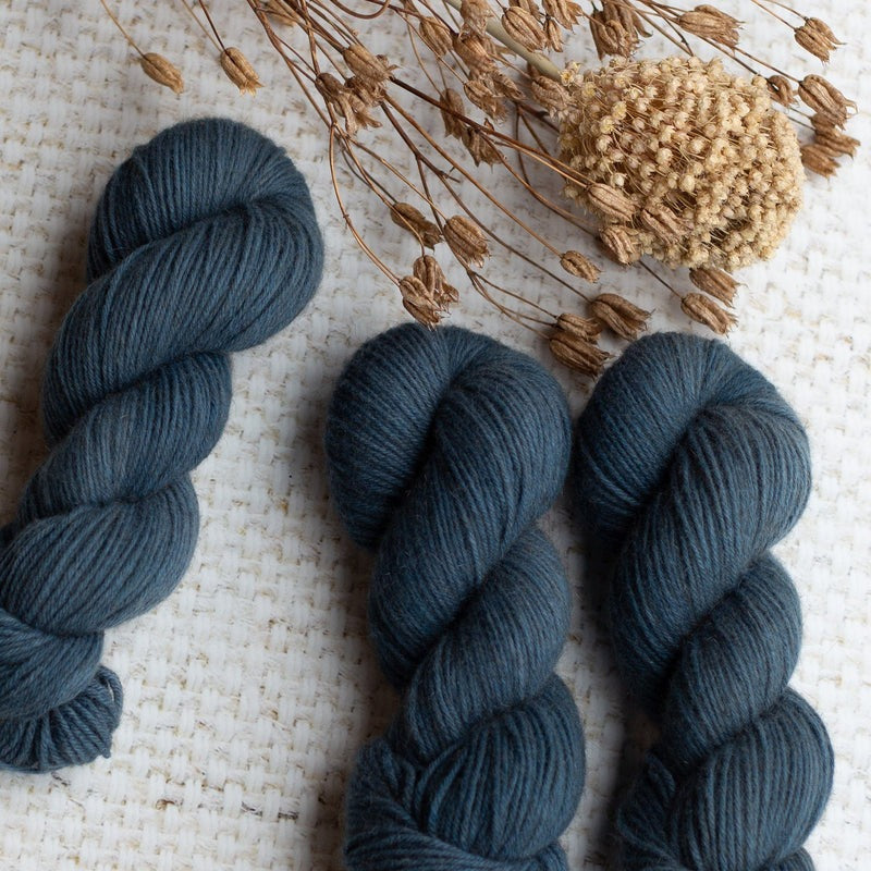 Three skeins of dark blue yarn with dried plant stems on a textured white surface.