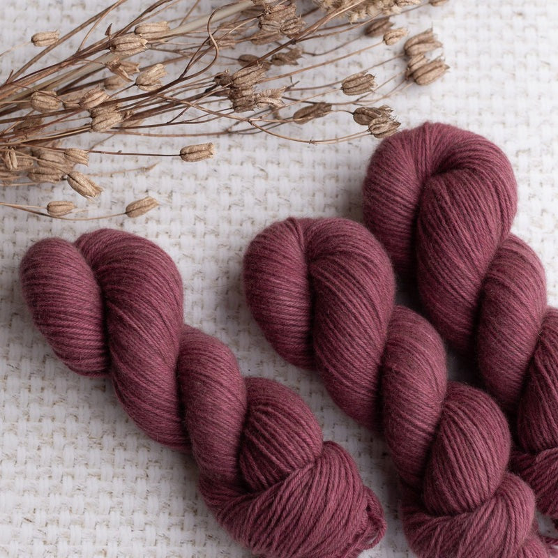 Four skeins of rich burgundy yarn on a textured white background with dried flowers for knitting and crochet projects.