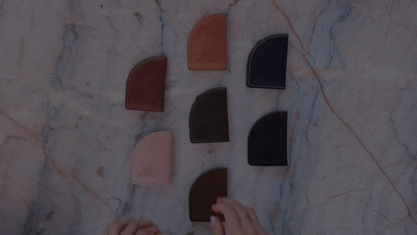 Six colorful leather wallet cardholders arranged on a marble surface with hands interacting.