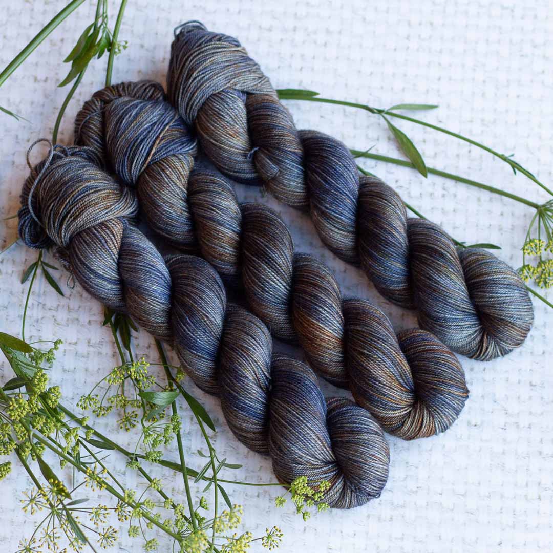 Four skeins of blue and brown variegated yarn on a white textured background with sprigs of green plants.