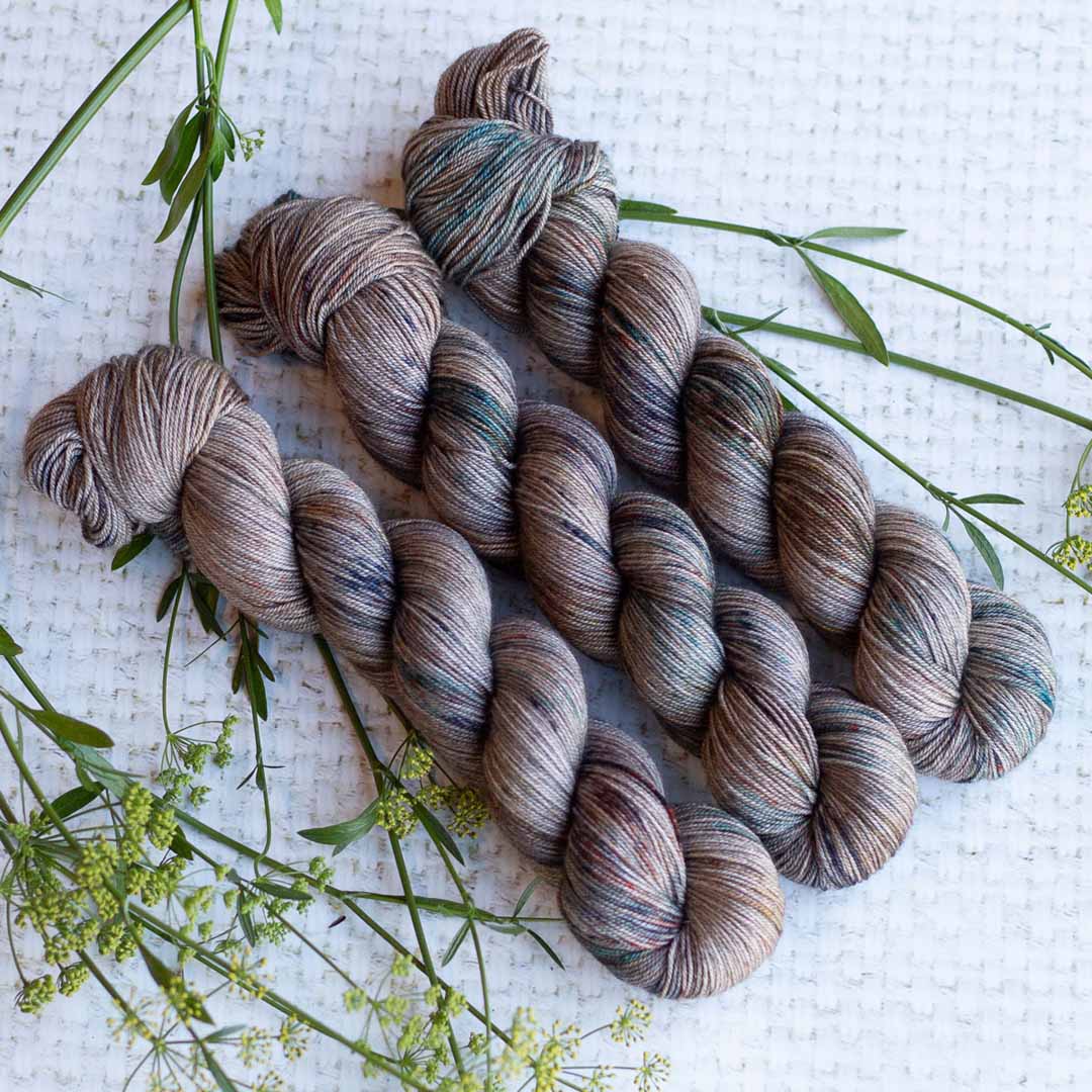Hand-dyed yarn skeins in earthy tones displayed on a soft white surface with green plant accents.