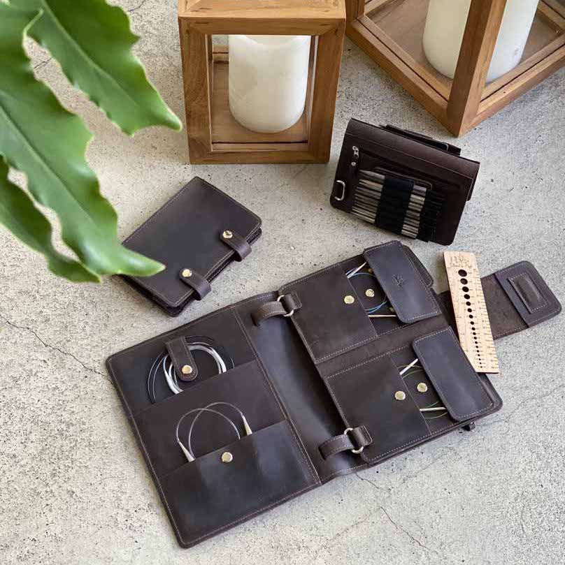 Leather knitting needle organizer set with circular needles, storage pouches, and measuring tool, displayed on a concrete surface near wooden candle holders and a green plant.