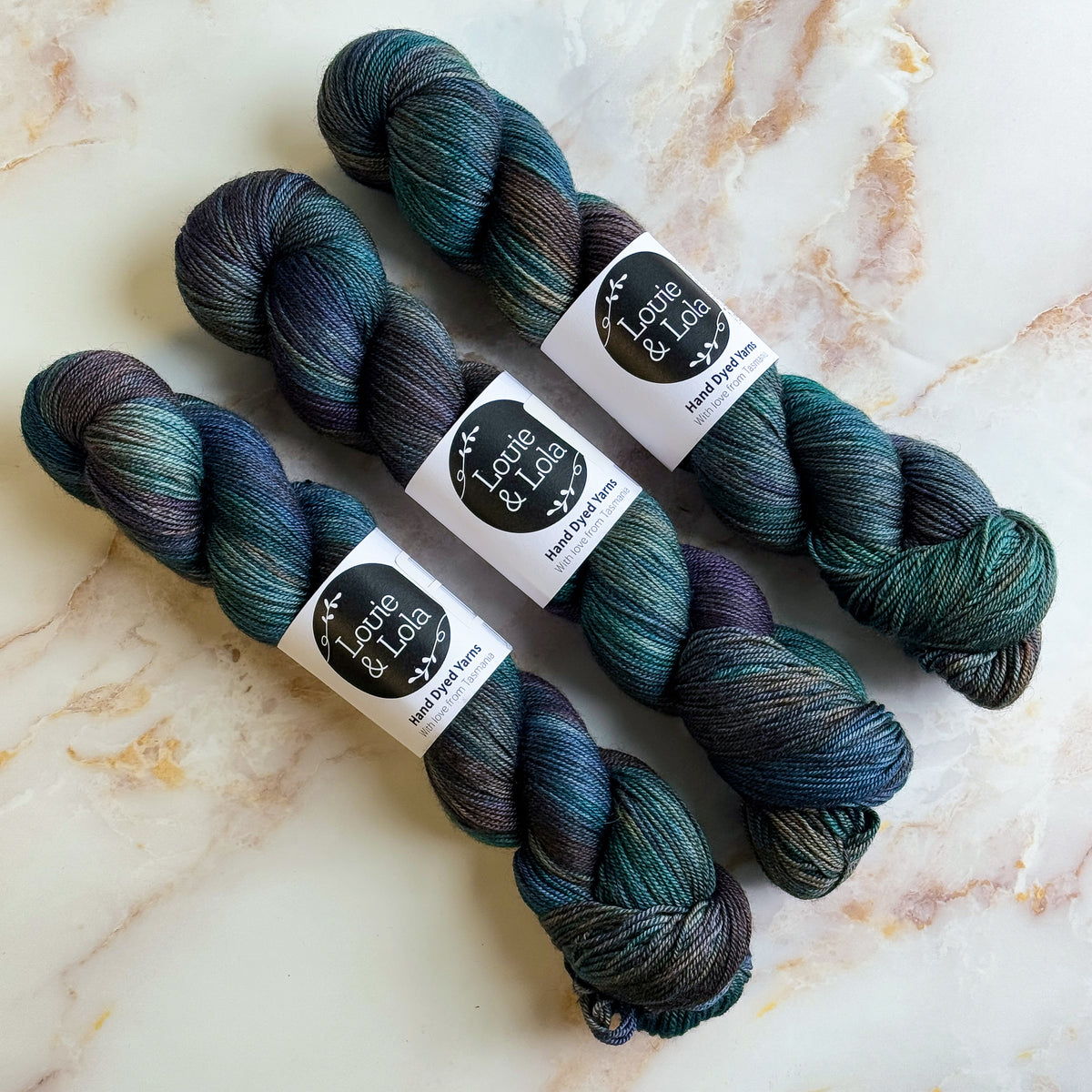 Three skeins of blue and green hand-dyed yarn on a marble surface.