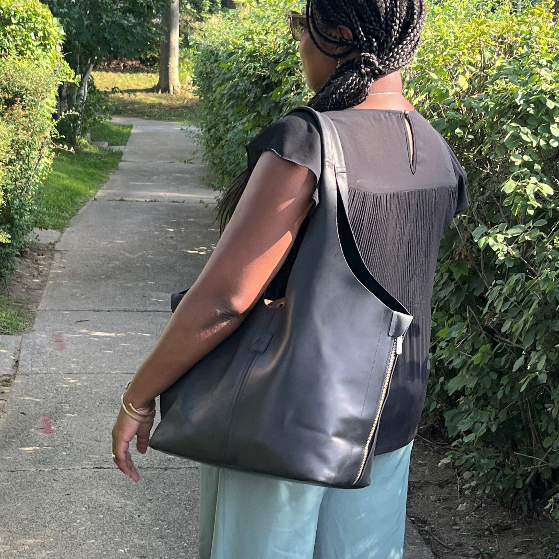 A person with braided hair wearing sunglasses and a black top, carrying a large black leather handbag while walking on a sidewalk next to green bushes.