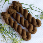 Three twisted skeins of brown yarn on a textured white background with sprigs of green foliage.