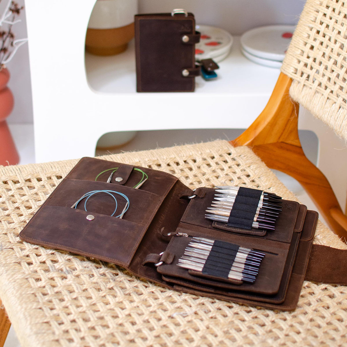 Open leather needle case displaying various sewing needles and colorful threads, placed on a woven chair in a cozy room.