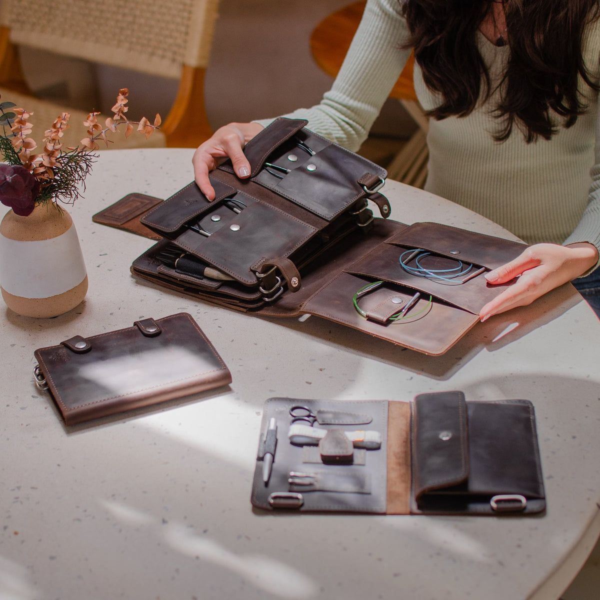 Person organizing leather planner and notebook on round table with decorative vase and chair in background.