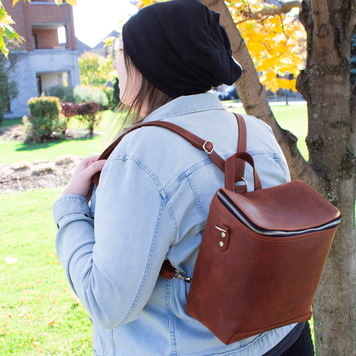 Woman wearing a denim jacket and a black beanie, carrying a brown leather backpack, standing next to a tree in a park during autumn.