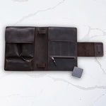 Dark brown leather travel organizer with multiple compartments, including a pen slot, on a light marble surface.