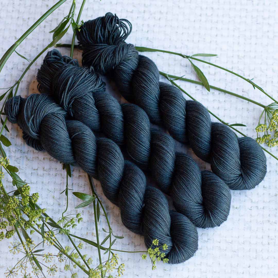 Four skeins of dark yarn on a textured white background with scattered green twigs and leaves.