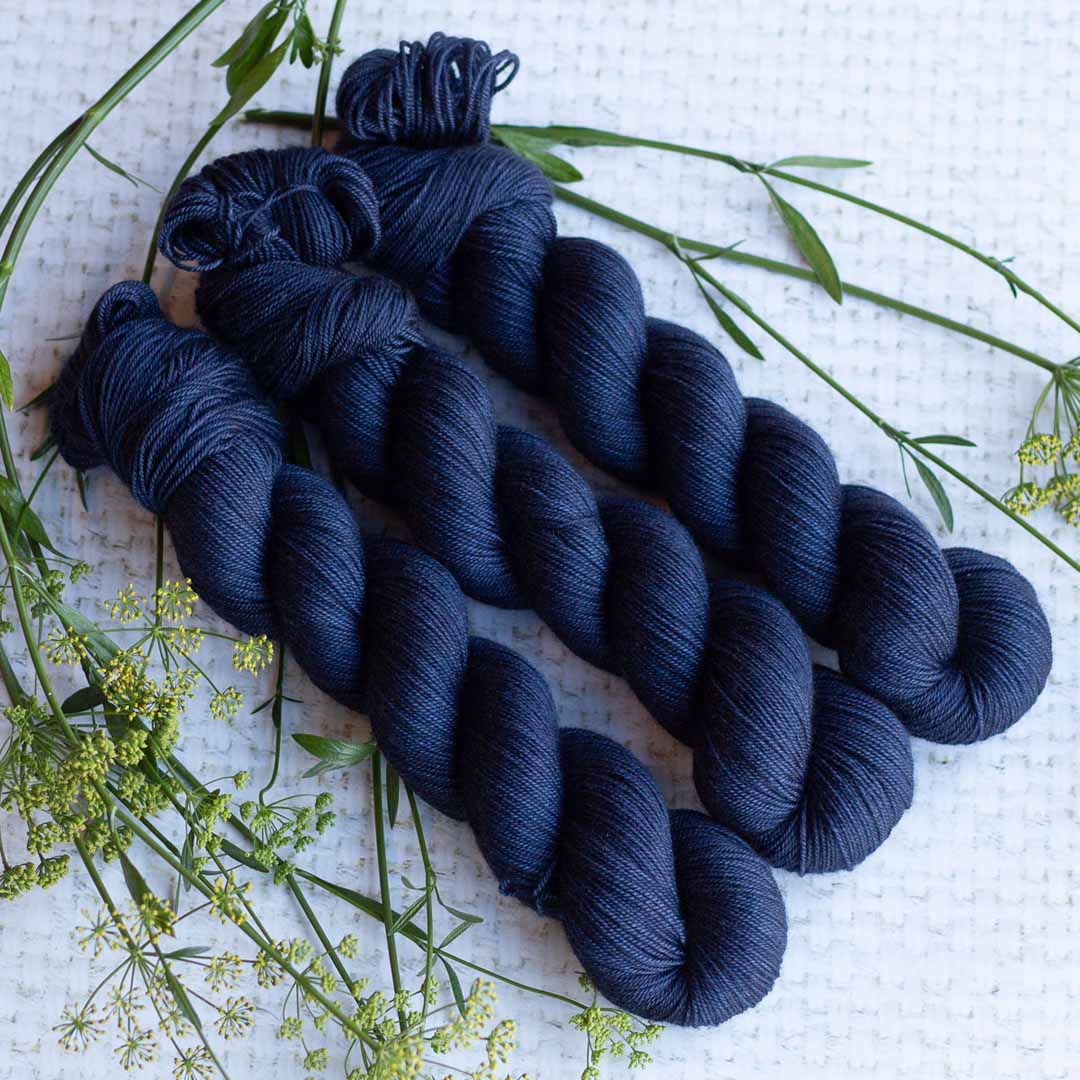 Three skeins of dark blue yarn arranged on a textured white surface with green foliage accents.