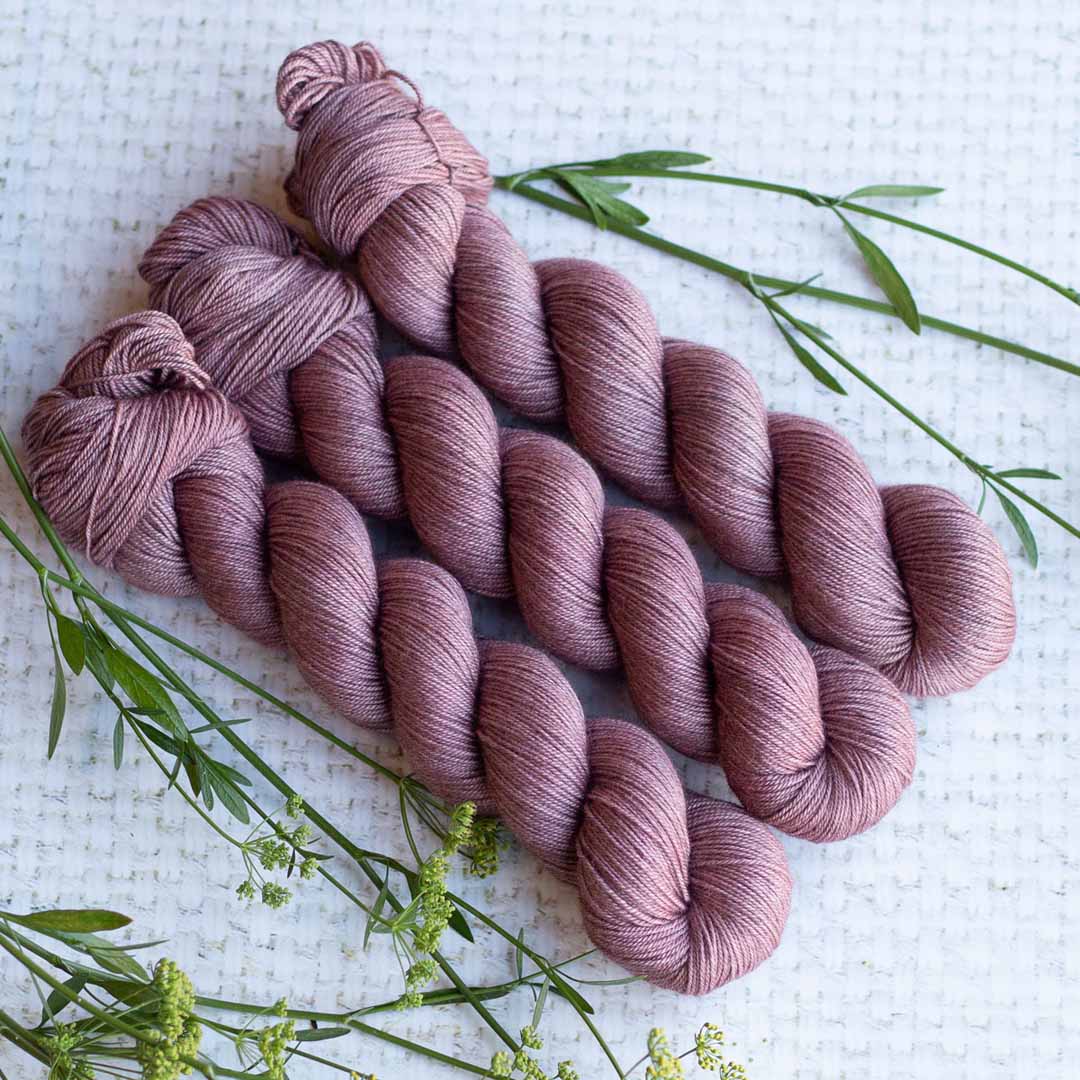 Three skeins of mauve-colored yarn with green foliage on a textured white background.