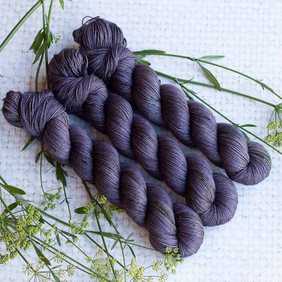 Four skeins of dark purple yarn arranged on a white textured surface with green foliage accents.