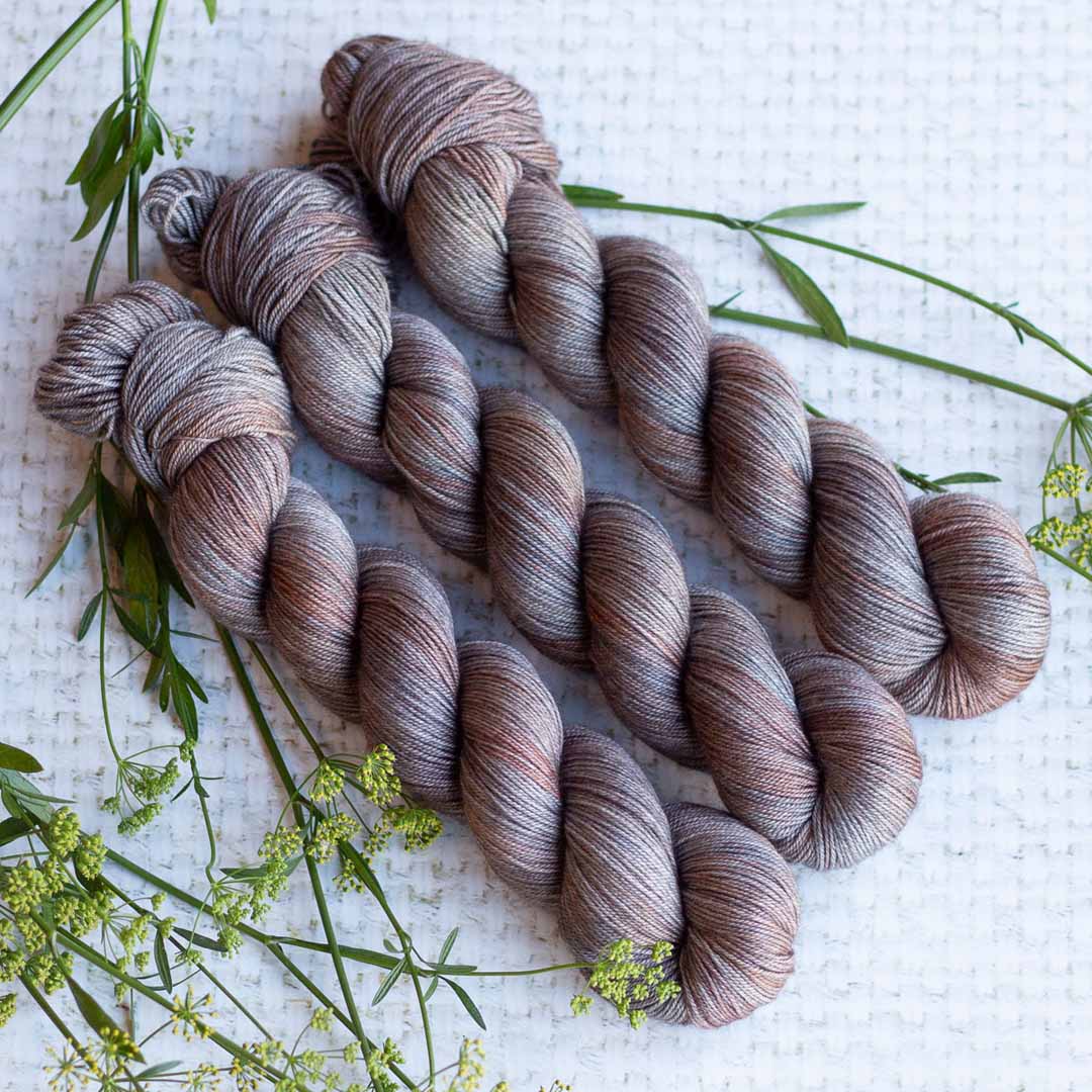 Three skeins of hand-dyed yarn in soft brown and gray tones on a textured white background with sprigs of green foliage.