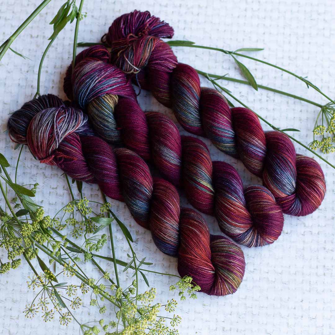 Hand-dyed skeins of colorful yarn with green foliage on a textured white background.
