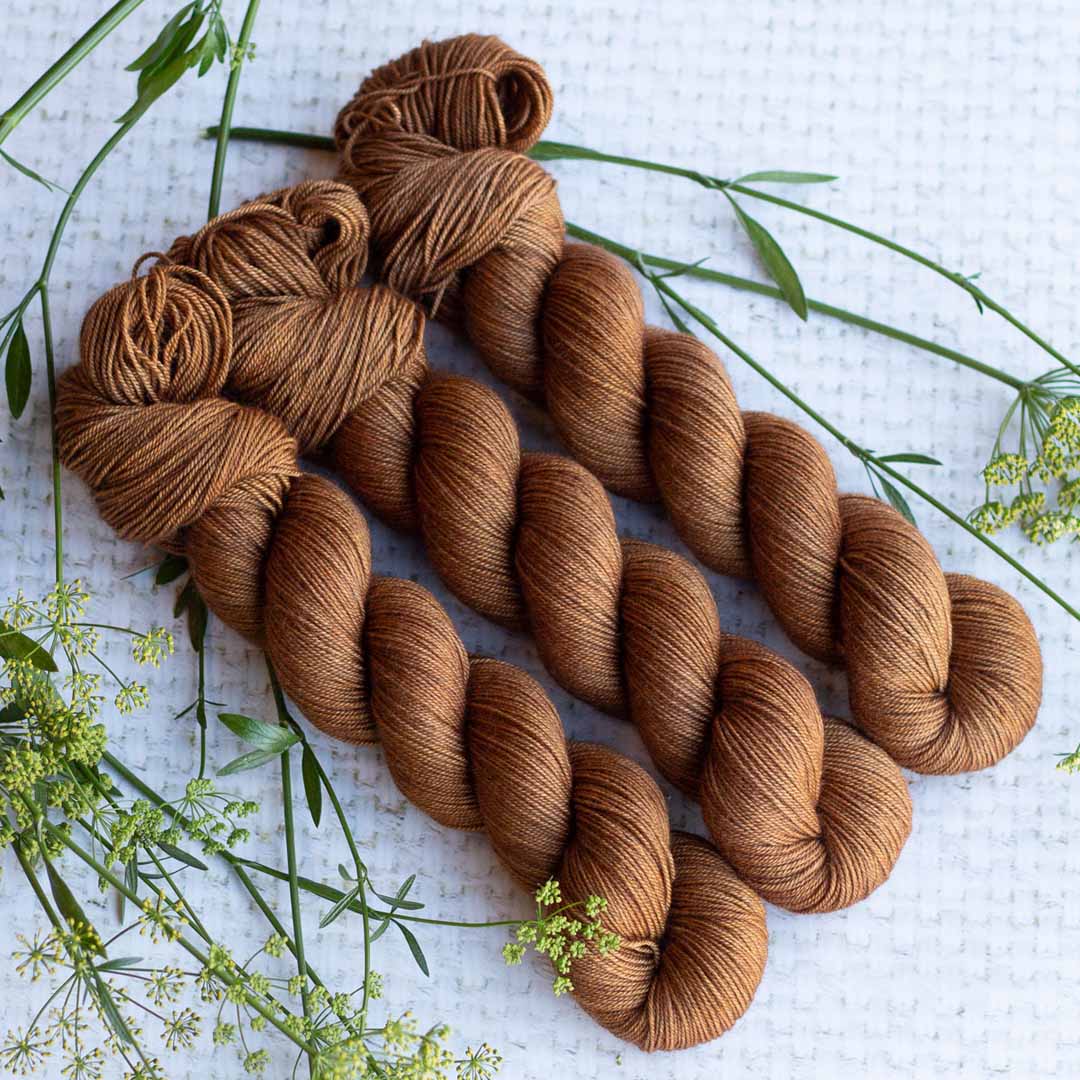 Three skeins of brown yarn on a textured white background with green plant sprigs.