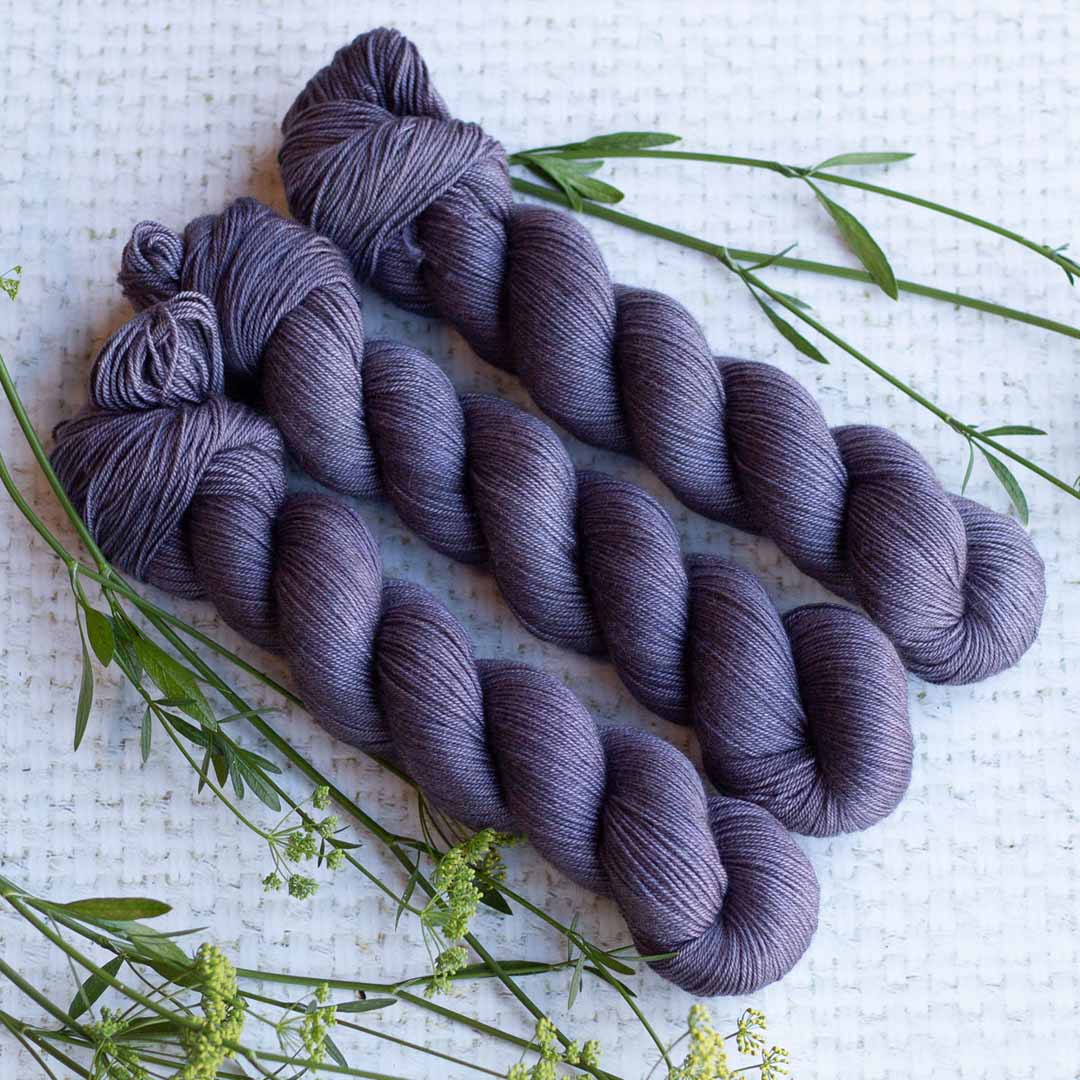 Three skeins of purple yarn on a textured white surface with green plant accents.