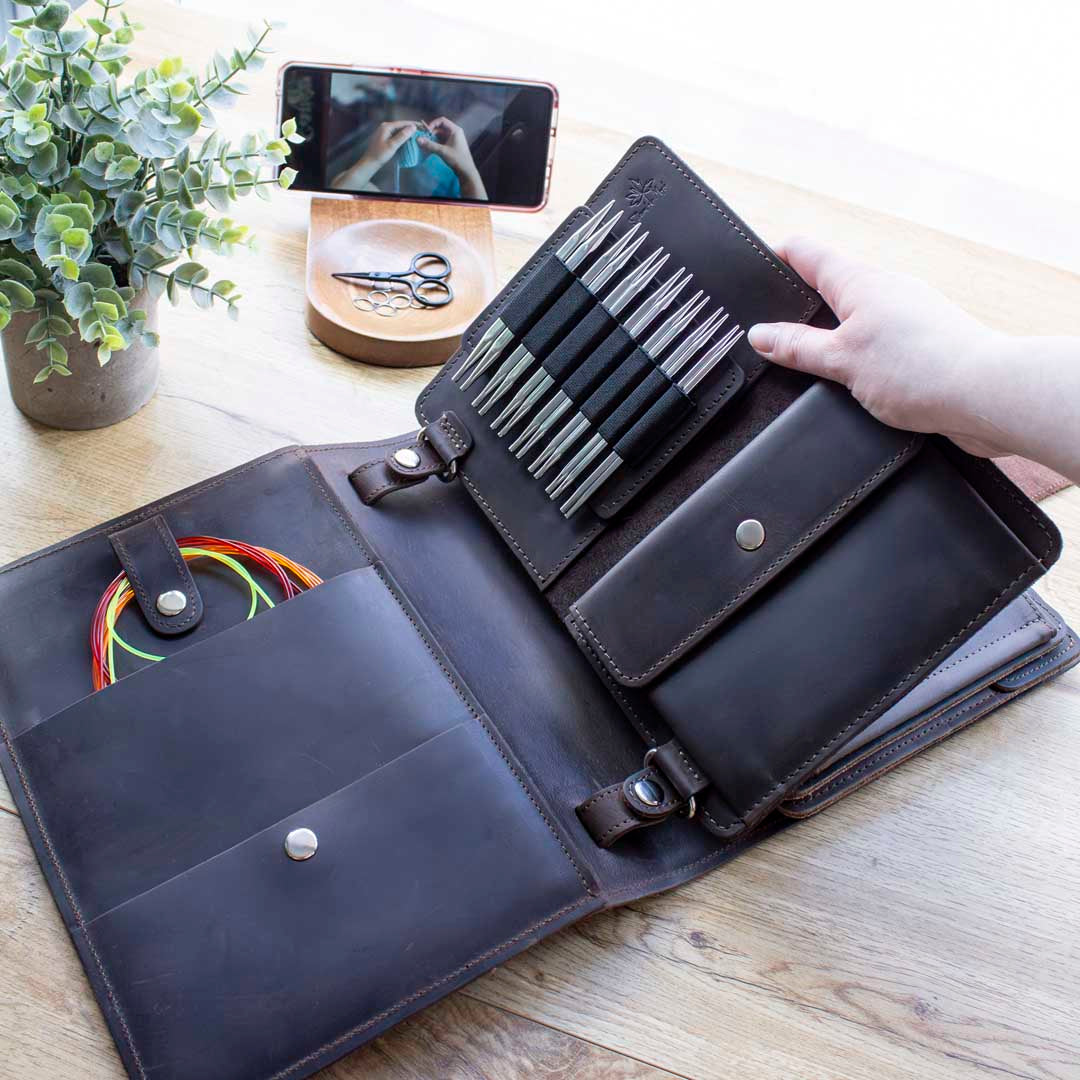 Leather knitting needle case open on a wooden table, containing various knitting tools including a set of knitting needles and colorful cables, with a smartphone in the background displaying a knitting tutorial video.