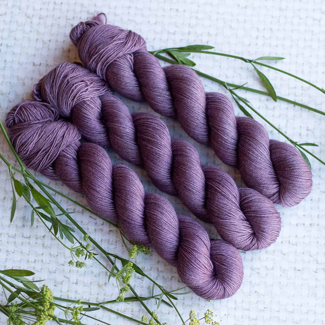 Three skeins of purple yarn on a textured white background with green sprigs.