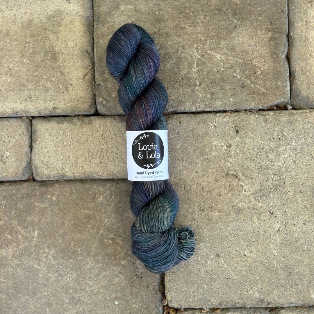 A skein of Louie & Lola hand dyed yarn in dark multicolored hues on a stone surface.