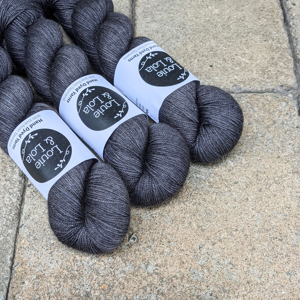 Three dark gray yarn skeins with labels arranged on a textured beige surface.