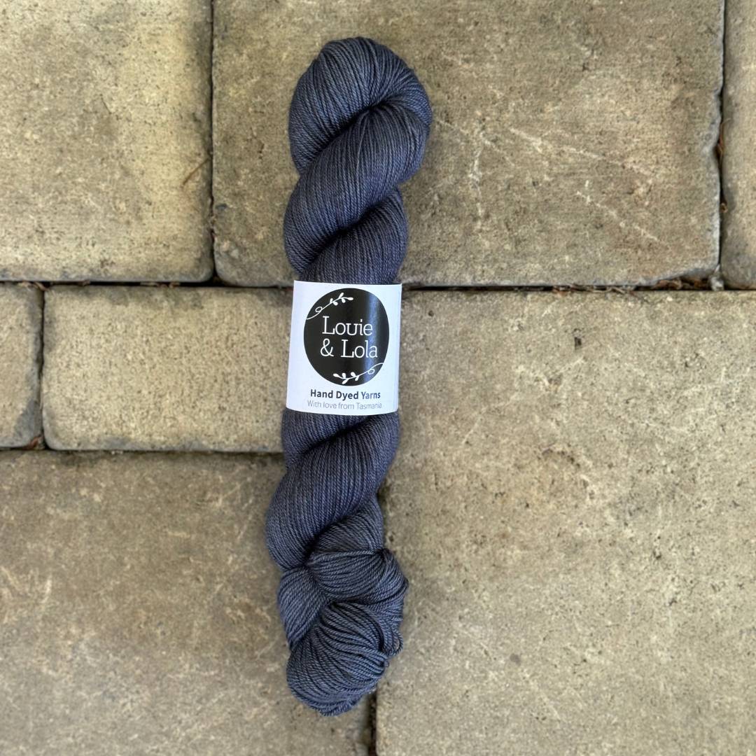A skein of dark gray Louie & Lola hand-dyed yarn on a textured stone background.