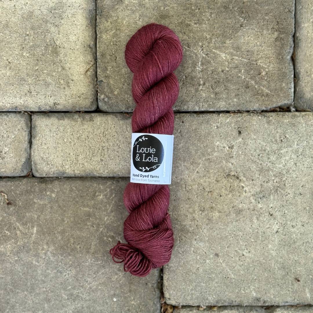 A skein of maroon colored yarn with a label, placed on a textured stone surface.