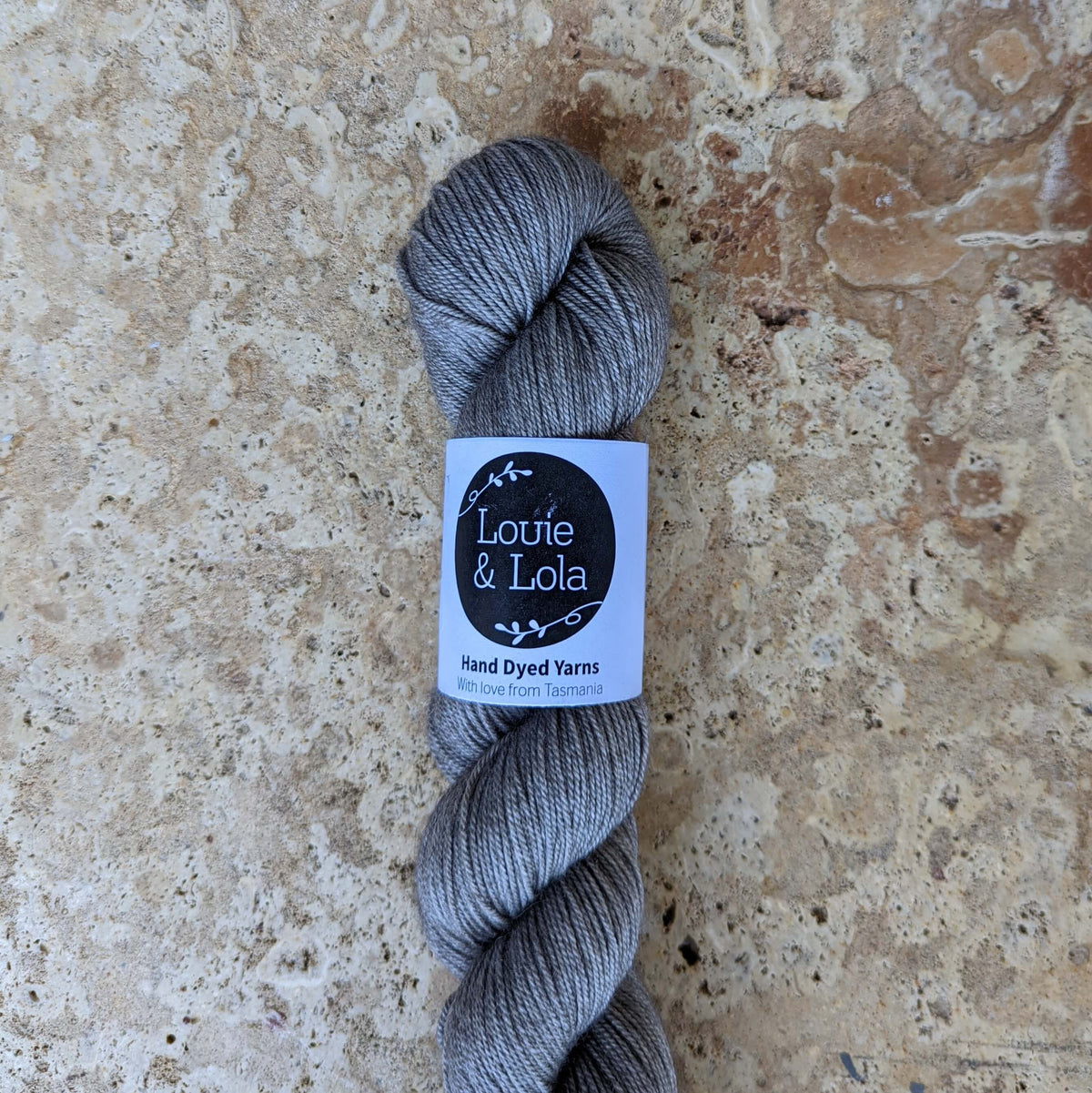 Gray skein of Luxurious Louie & Lola Fingering Yarn on textured background.