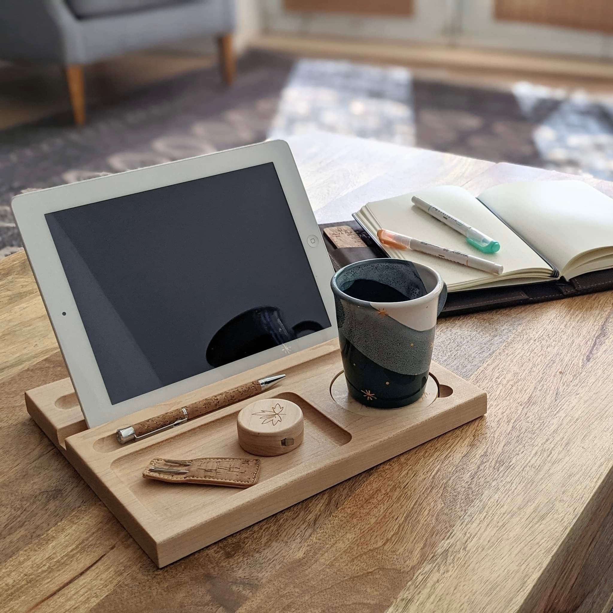 Tablet on a wooden stand with a ceramic mug, pen, small wooden case, and open notebook with markers on a wooden desk.
