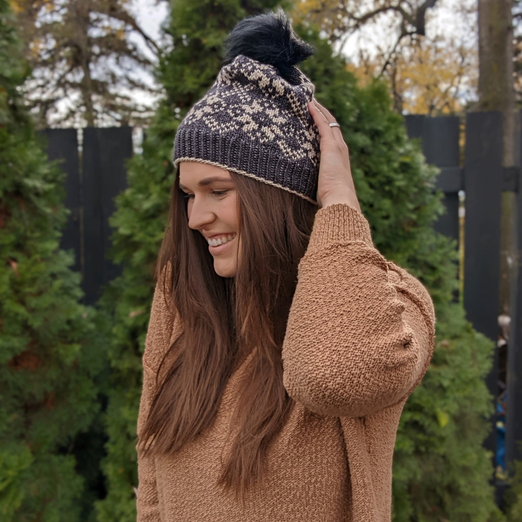 Woman with long hair wearing a knitted beanie and sweater in a garden with green trees.