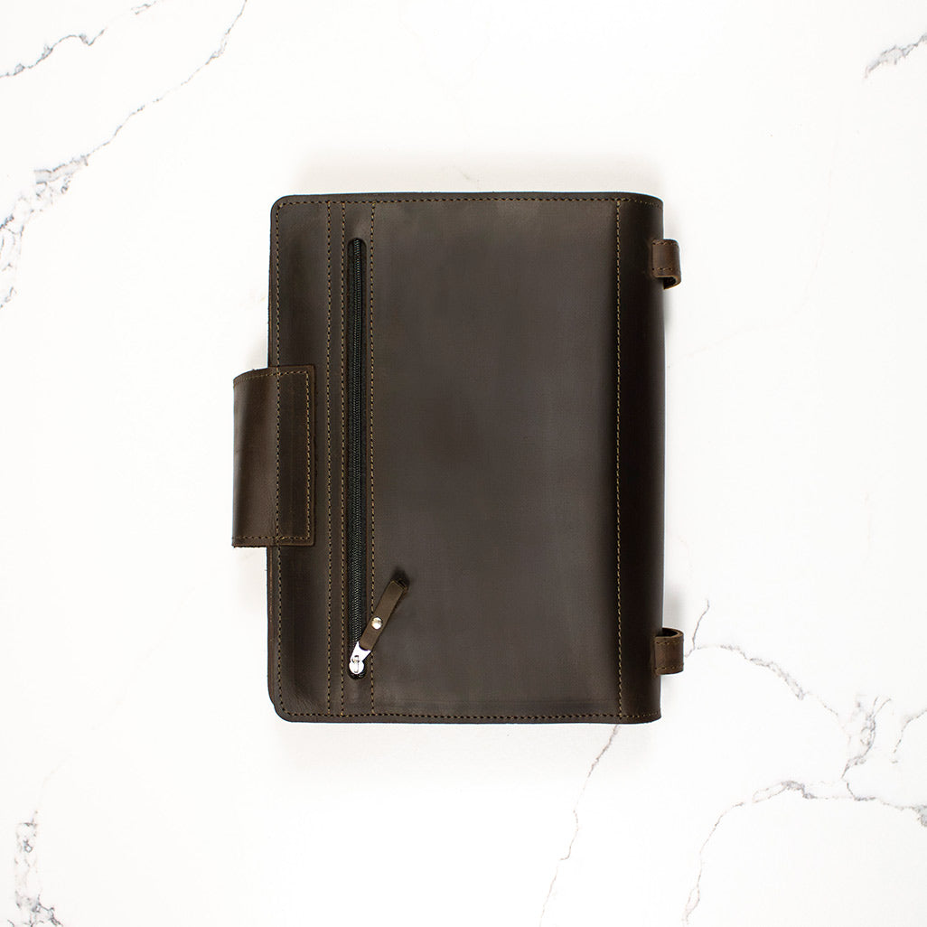 Dark brown leather notebook cover on a marble background with a pen inside a zippered pocket.