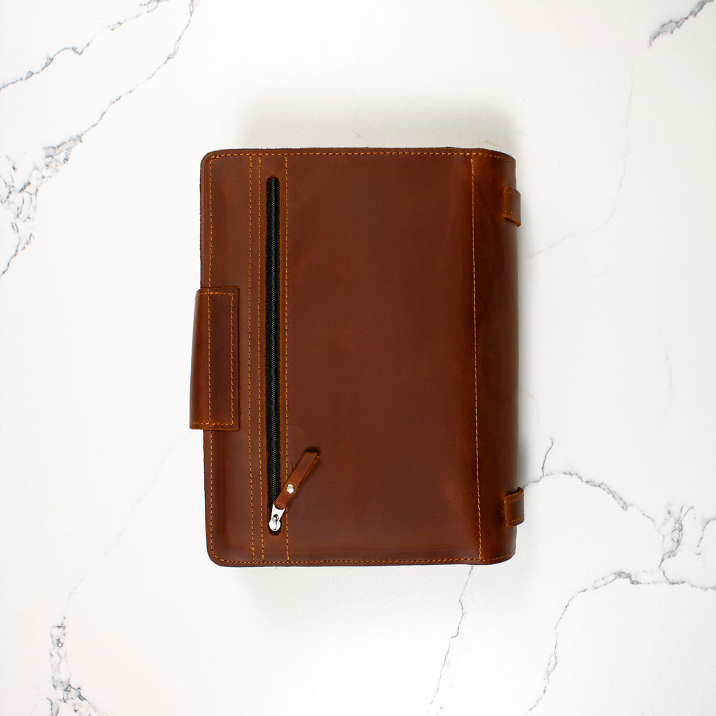 Brown leather binder with a zipper pocket on a marble surface.