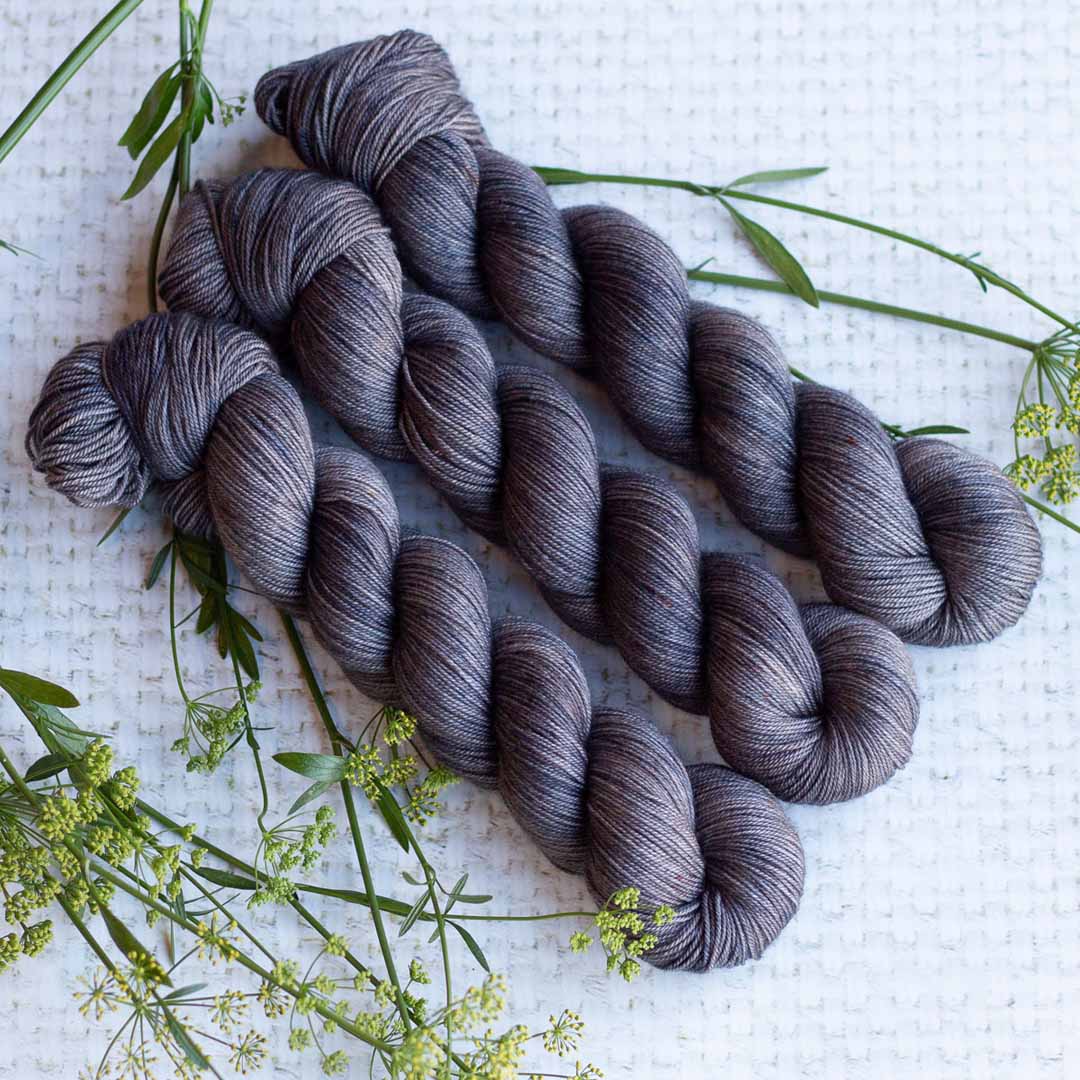 Three skeins of gray yarn arranged on a textured white surface with green plants for knitting projects.