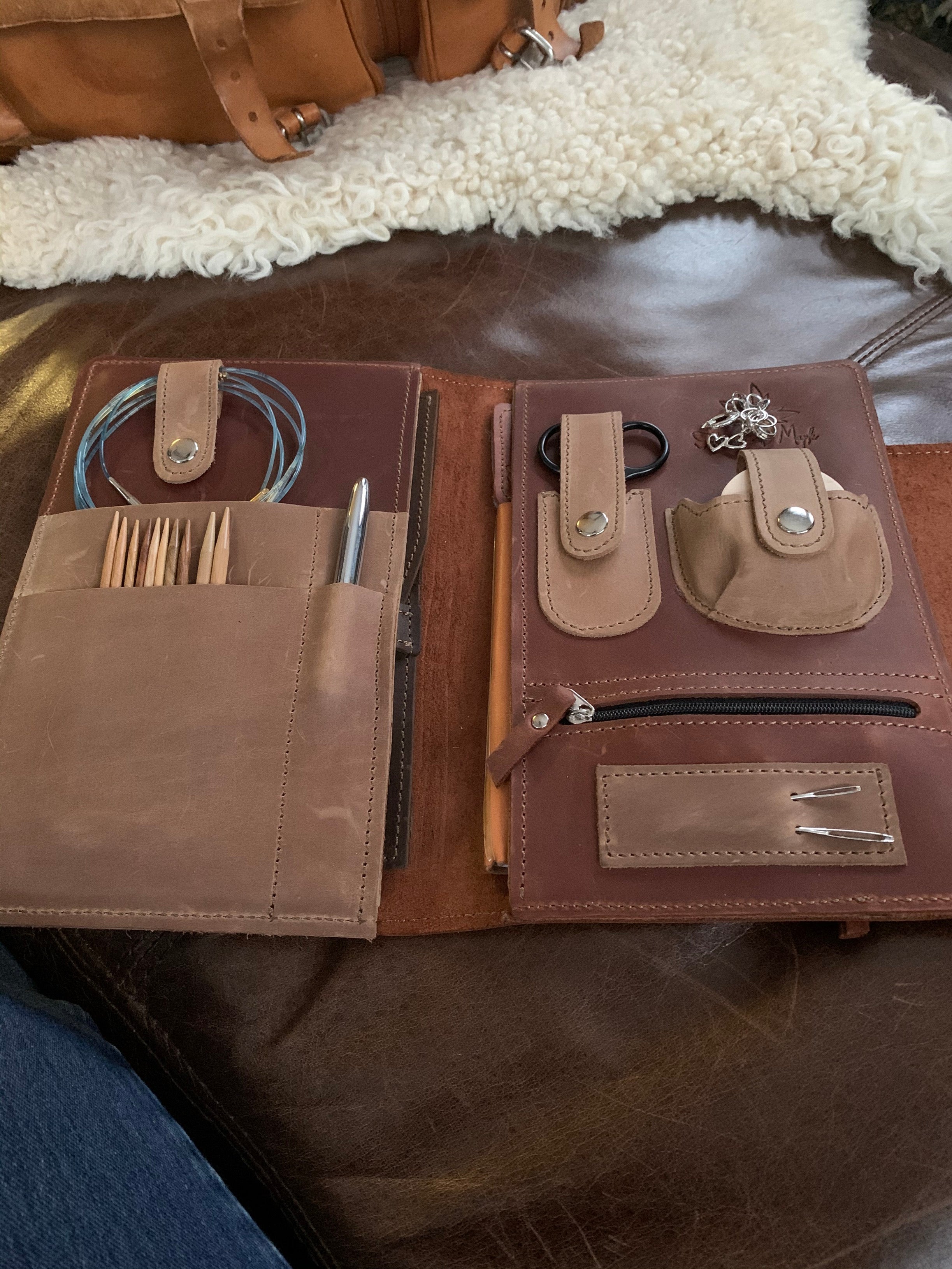 Leather knitting needle holder set with circular needles, wooden knitting needles, metal loops, and pen on a leather couch.