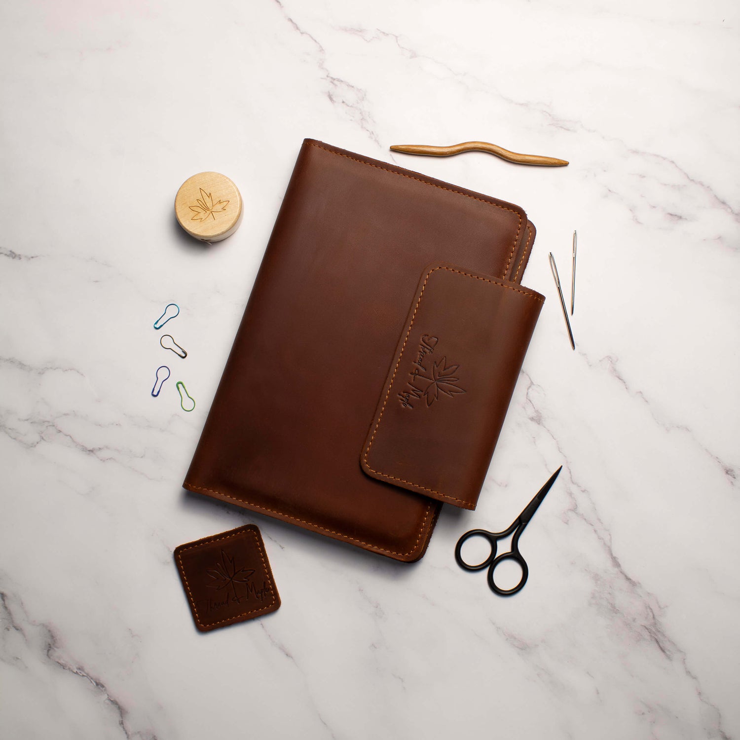 Brown leather notebook and accessories on a marble surface with sewing tools and a pair of scissors.