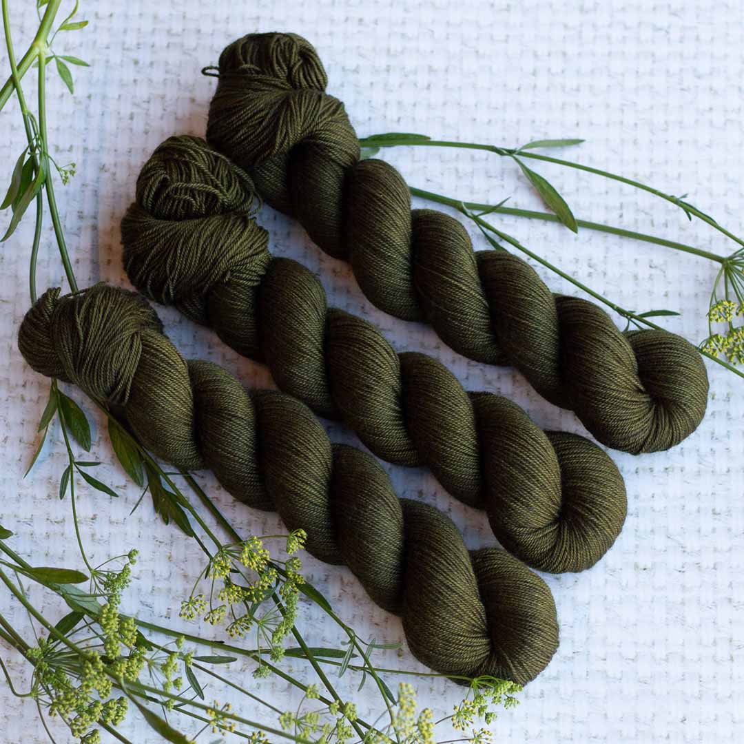 Three skeins of dark green yarn on a white textured surface with sprigs of greenery.