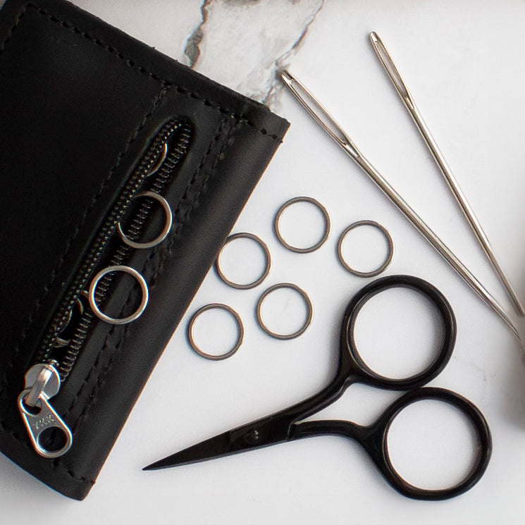 Black leather wallet with zipper, silver knitting needles, small black scissors, and metal stitch markers on a white surface.