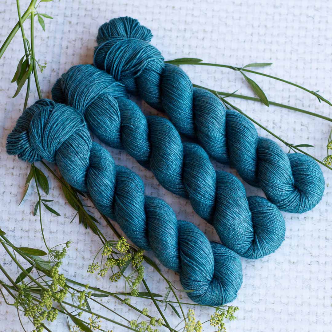 Three skeins of teal yarn arranged on a textured white surface with greenery accents.
