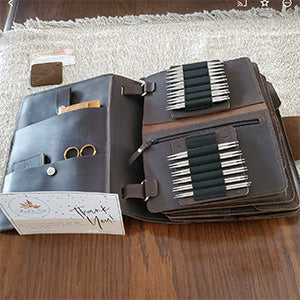 Leather knitting needle organizer open on a wooden table, showcasing various knitting needles and accessories.