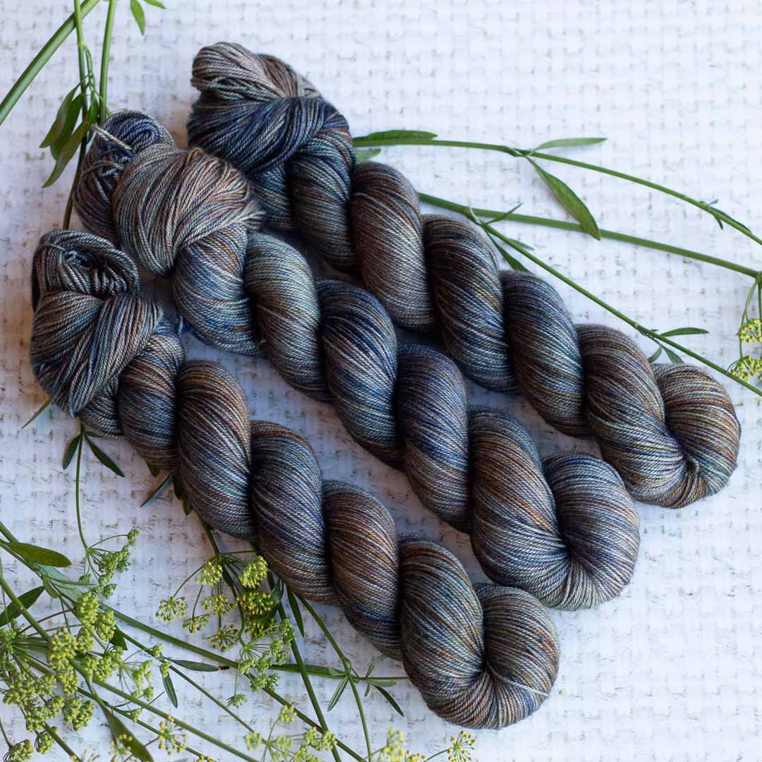 Three skeins of blue and grey hand-dyed yarn arranged on a white surface with greenery accents.